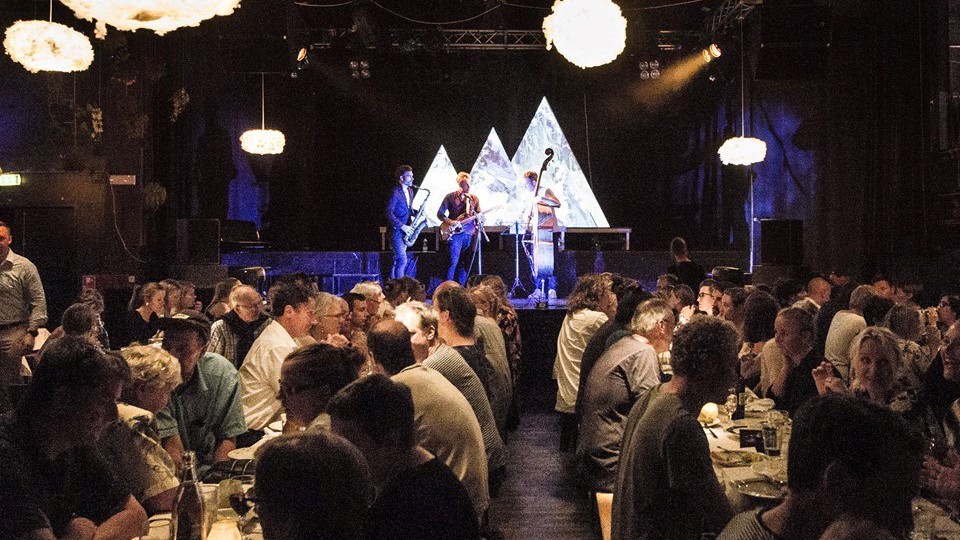 Mennesker siddende ved langborde, der spiser og hygger sig. I baggrunden på en scene står tre musikere og spiller.